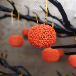 K&ouml;nnte beinhalten: Orangefarbene, runde, spitzenartige Ornamente, die an schwarzen &Auml;sten h&auml;ngen. Die Ornamente bestehen aus einem zarten, gewebten Material und haben eine leicht strukturierte Oberfl&auml;che.