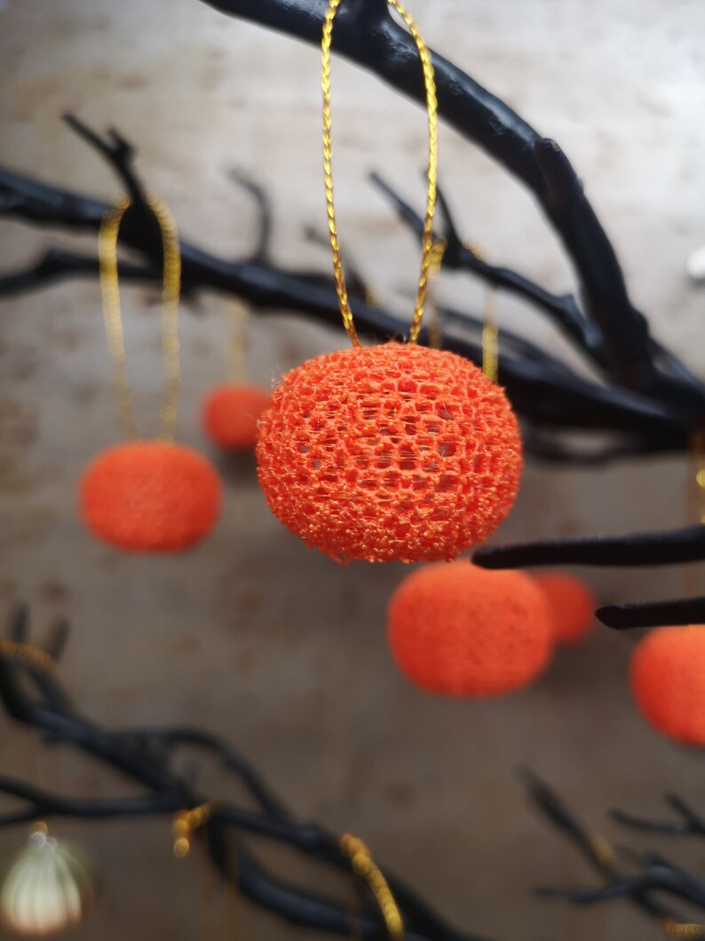 K&ouml;nnte beinhalten: Orangefarbene, runde, spitzenartige Ornamente, die an schwarzen &Auml;sten h&auml;ngen. Die Ornamente bestehen aus einem zarten, gewebten Material und haben eine leicht strukturierte Oberfl&auml;che.