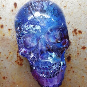 May include: A blue and purple glass skull with white swirls and a galaxy-like design. The skull is sitting on a rusty metal surface.