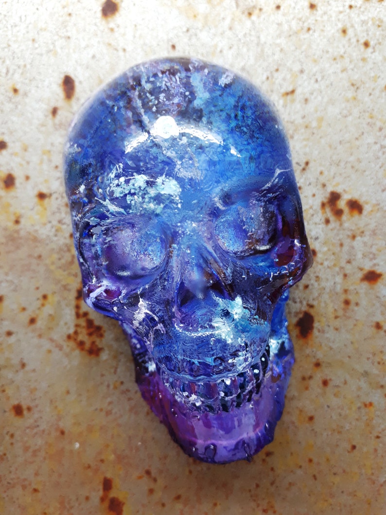May include: A blue and purple glass skull with white swirls and a galaxy-like design. The skull is sitting on a rusty metal surface.
