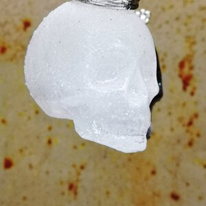 May include: A white, sparkly skull pendant with a silver crown on top. The pendant is hanging from a silver chain.