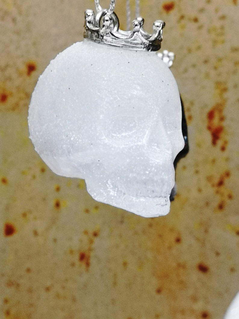 May include: A white, sparkly skull pendant with a silver crown on top. The pendant is hanging from a silver chain.