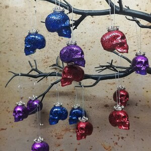 May include: A collection of 12 glittery skull ornaments with silver crowns hanging from a branch. The skulls are in three colors: blue, red, and purple.