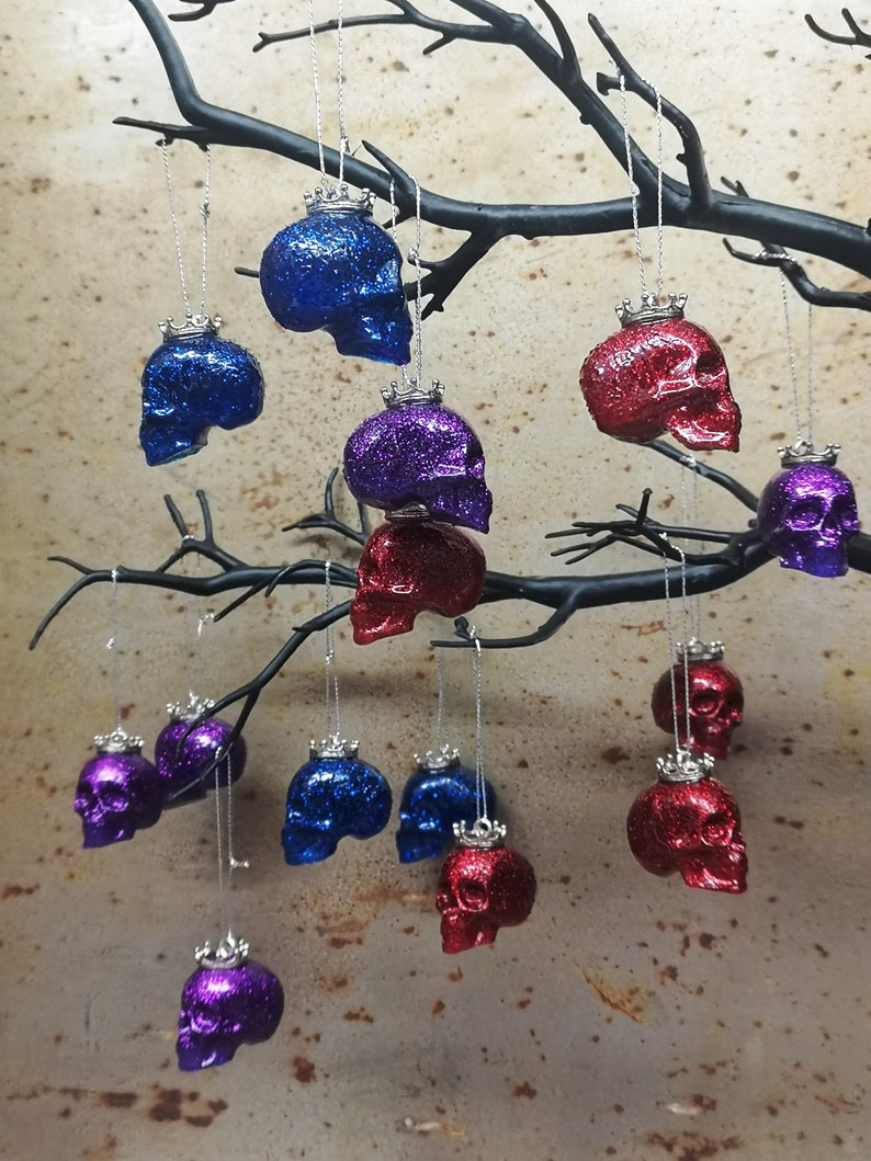 May include: A collection of 12 glittery skull ornaments with silver crowns hanging from a branch. The skulls are in three colors: blue, red, and purple.
