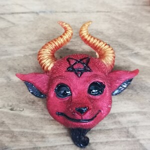 Puede incluir: Una figurita roja con forma de diablo con cuernos dorados y un pentagrama negro en la frente. La figurita tiene ojos negros y una peque&ntilde;a barba negra.