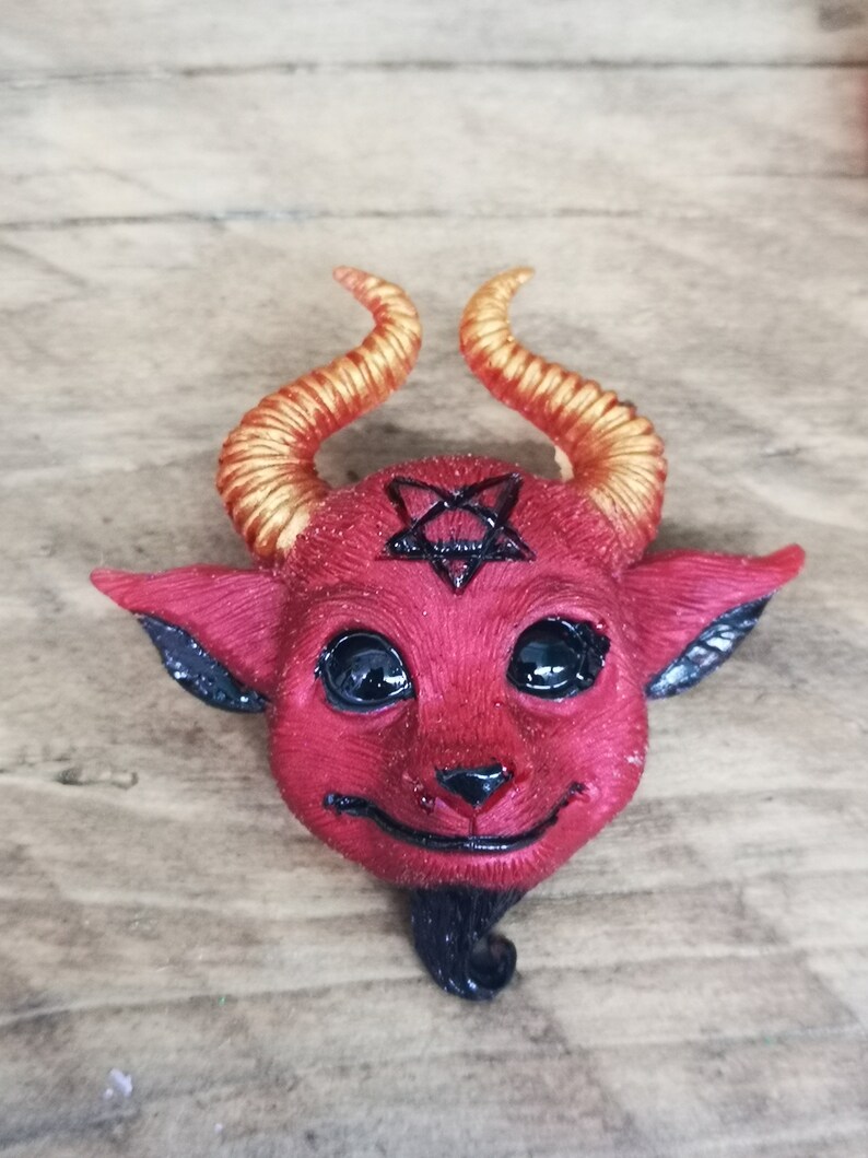 Puede incluir: Una figurita roja con forma de diablo con cuernos dorados y un pentagrama negro en la frente. La figurita tiene ojos negros y una peque&ntilde;a barba negra.
