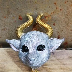 Puede incluir: Una figurita de color plateado de una criatura con ojos grandes, nariz puntiaguda y orejas peque&ntilde;as. La criatura tiene dos cuernos dorados y un s&iacute;mbolo de pentagrama en la frente.
