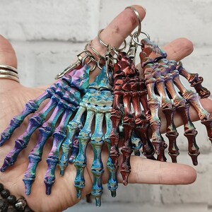 May include: A set of five skeleton hand keychains with iridescent finishes in shades of blue, purple, and copper. The keychains are made of a durable material and feature intricate details.