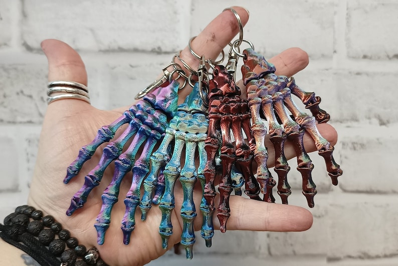 May include: A set of five skeleton hand keychains with iridescent finishes in shades of blue, purple, and copper. The keychains are made of a durable material and feature intricate details.