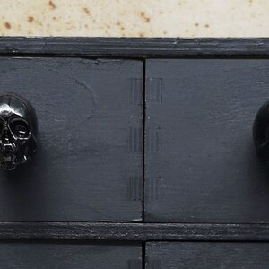 May include: Black wooden chest of drawers with four drawers. Each drawer has a black skull-shaped knob.