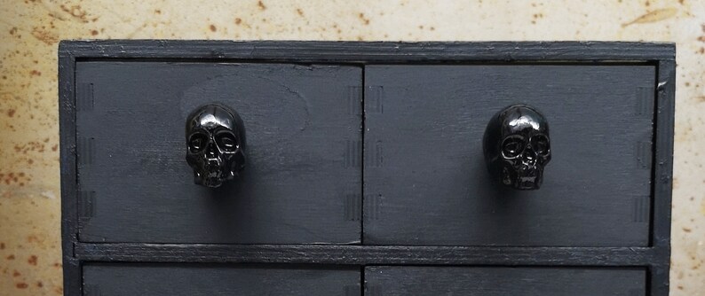 May include: Black wooden chest of drawers with four drawers. Each drawer has a black skull-shaped knob.