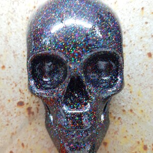 May include: A black skull with iridescent glitter covering the surface. The skull has a detailed design with eye sockets, a nose, and a mouth.