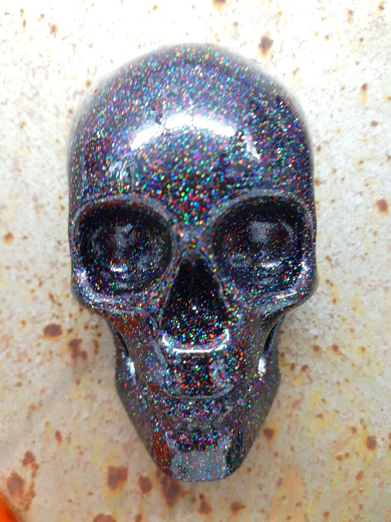 May include: A black skull with iridescent glitter covering the surface. The skull has a detailed design with eye sockets, a nose, and a mouth.