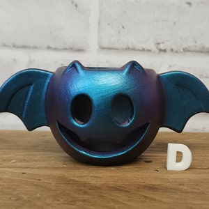 May include: A teal and purple iridescent ceramic bat planter with a smiling face. The planter is sitting on a wooden surface.