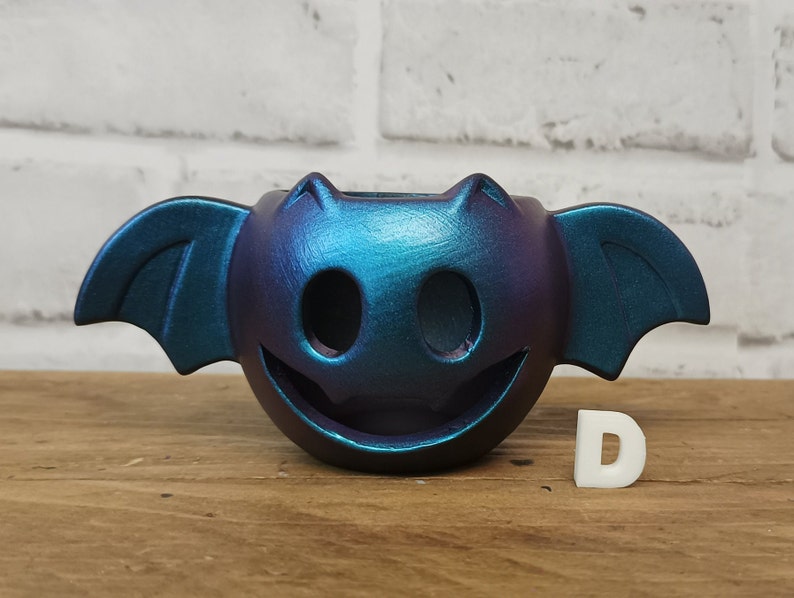 May include: A teal and purple iridescent ceramic bat planter with a smiling face. The planter is sitting on a wooden surface.