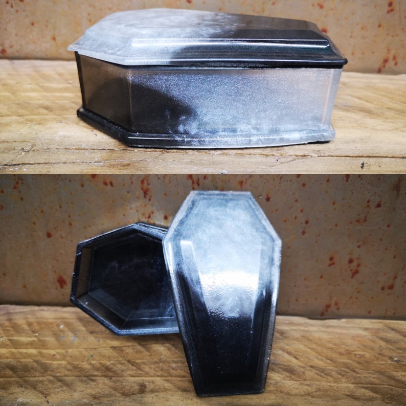 May include: A black and white coffin-shaped box with a lid. The box is made of resin and has a smooth, glossy finish. The lid is slightly ajar, revealing the interior of the box.