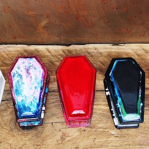 May include: A set of eight small coffin-shaped boxes in various colors, including purple, white, blue, red, black, and gray. The boxes are made of resin and have a glossy finish.