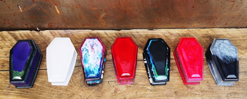 May include: A set of eight small coffin-shaped boxes in various colors, including purple, white, blue, red, black, and gray. The boxes are made of resin and have a glossy finish.