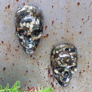 May include: Two small resin skull sculptures with a textured surface made of small stones in shades of brown, white, and black.