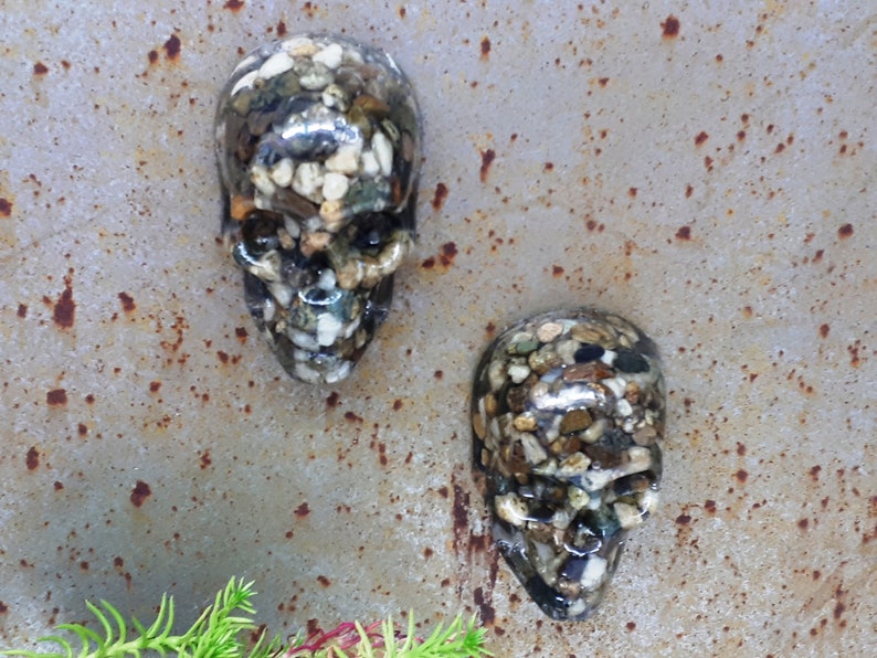 May include: Two small resin skull sculptures with a textured surface made of small stones in shades of brown, white, and black.