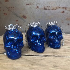 May include: Three blue glitter skull ornaments with silver crowns. Each skull has a silver loop for hanging.