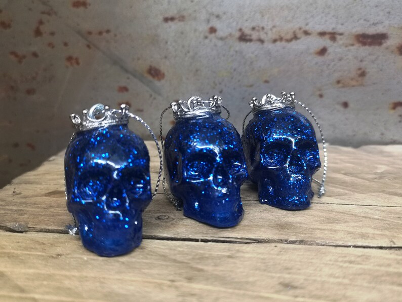May include: Three blue glitter skull ornaments with silver crowns. Each skull has a silver loop for hanging.