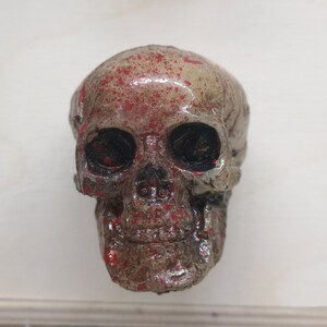 May include: A small, white skull with red paint splatters. The skull has black eye sockets and a detailed jawline.