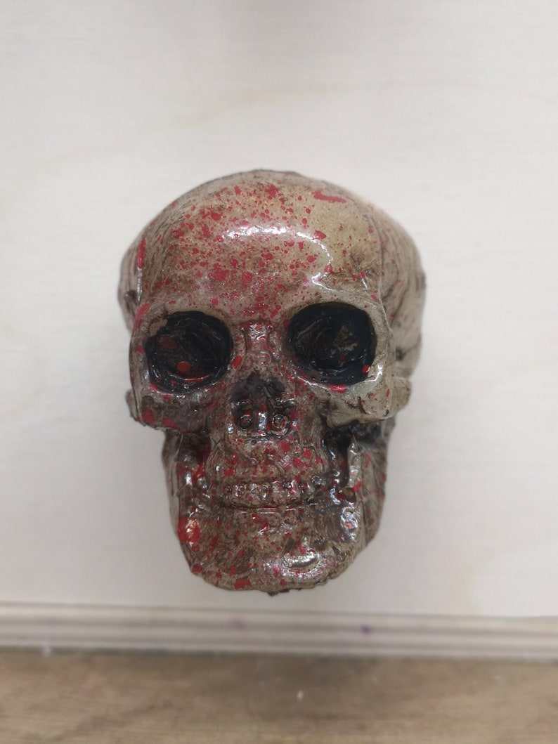May include: A small, white skull with red paint splatters. The skull has black eye sockets and a detailed jawline.