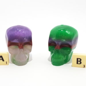 May include: Two small resin skull figurines, one with a purple and green color scheme and the other with a green and purple color scheme. The skulls are sitting on a white surface with letter tiles that say "A1" and "B3".