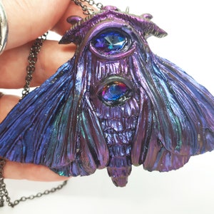 May include: A dark purple and blue iridescent moth pendant necklace with two large blue gemstone eyes. The necklace has a black chain.