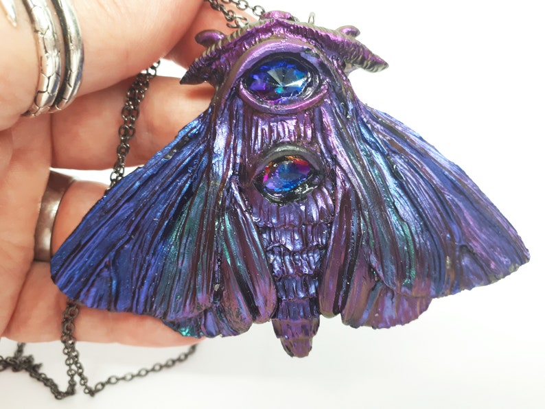 May include: A dark purple and blue iridescent moth pendant necklace with two large blue gemstone eyes. The necklace has a black chain.