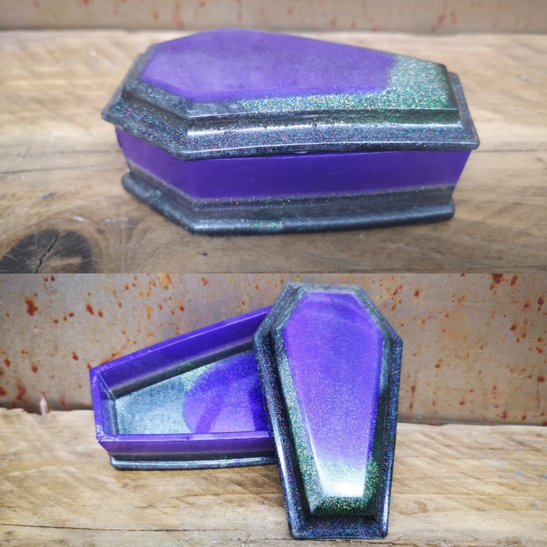 May include: A purple and black coffin-shaped trinket box with a glittery finish. The box is open and the lid is laying next to it.