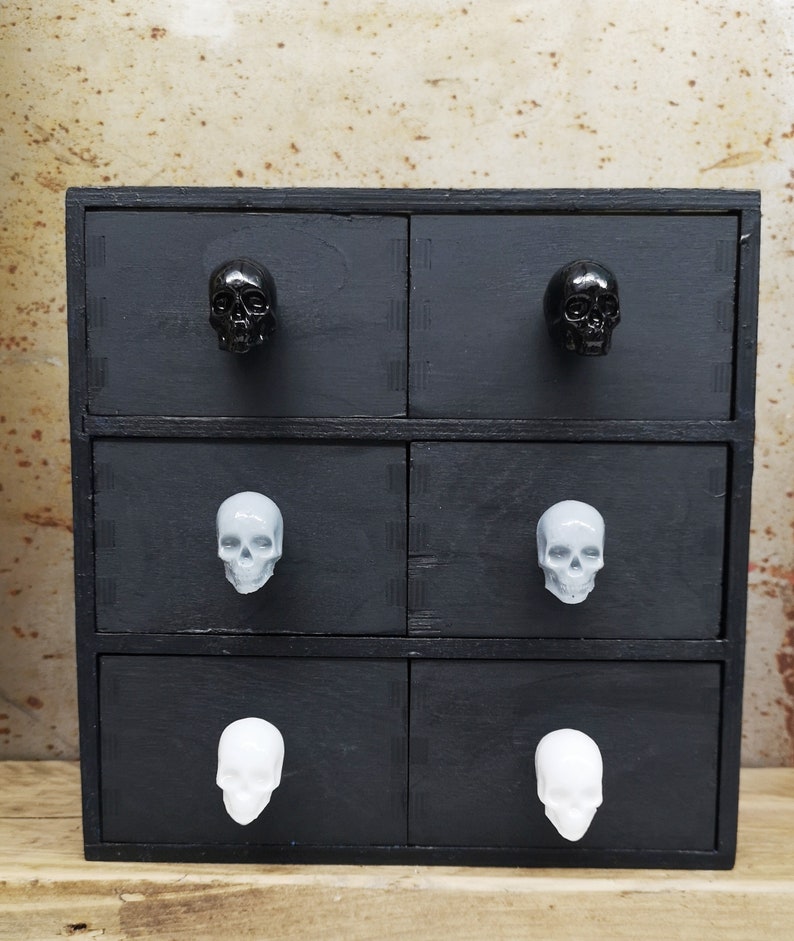 May include: A black wooden chest of drawers with six drawers. Each drawer has a skull-shaped knob. The top two drawers have black skull knobs, the middle two drawers have grey skull knobs, and the bottom two drawers have white skull knobs.