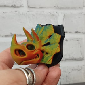 Puede incluir: Una cabeza de triceratops impresa en 3D en tonos naranja, amarillo y azul, montada sobre una base negra. La cabeza del dinosaurio presenta texturas detalladas y una expresión juguetona, mostrando un gradiente de color vibrante.