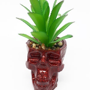 Pode incluir: Um vaso de caveira vermelho com glitter com uma planta suculenta verde dentro. O vaso &eacute; feito de cer&acirc;mica e tem um acabamento brilhante.