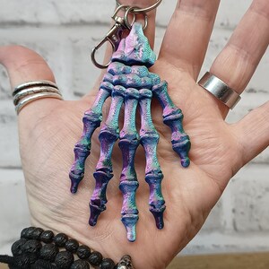 May include: A skeleton hand keyring with a blue, purple and green iridescent finish. The keyring is attached to a silver metal ring.