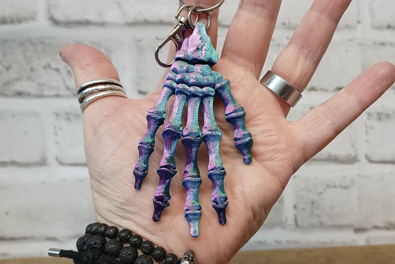 May include: A skeleton hand keyring with a blue, purple and green iridescent finish. The keyring is attached to a silver metal ring.