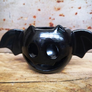 May include: Black ceramic bat candle holder with cut-out eyes and a smiling mouth. The holder has wings on either side.