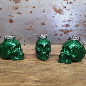 May include: Three green glitter skull ornaments with silver crowns. Each skull has a silver loop for hanging.