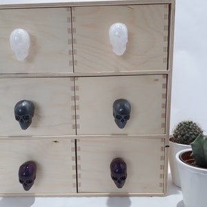 May include: A set of six wooden drawers with skull-shaped drawer pulls. The drawers are arranged in two stacks of three. The top two drawers have white skull pulls, the middle two drawers have gray skull pulls, and the bottom two drawers have purple skull pulls.