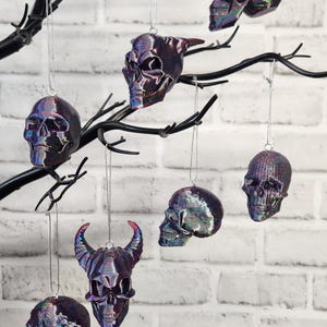May include: Seven iridescent skull ornaments hanging from a black branch. The skulls are various shades of purple, blue, and green. The ornaments are hanging against a white brick wall.