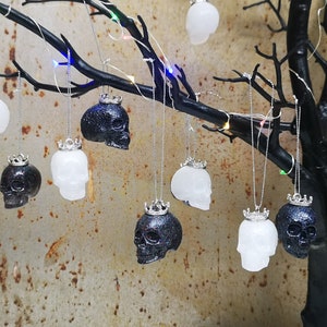 May include: A collection of black and white skull ornaments with silver crowns hanging from a branch. The ornaments are made of resin and have a glittery finish.