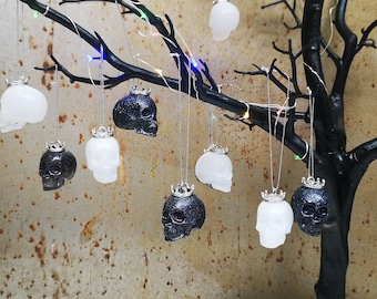 May include: A collection of black and white skull ornaments with silver crowns hanging from a branch. The ornaments are made of resin and have a glittery finish.