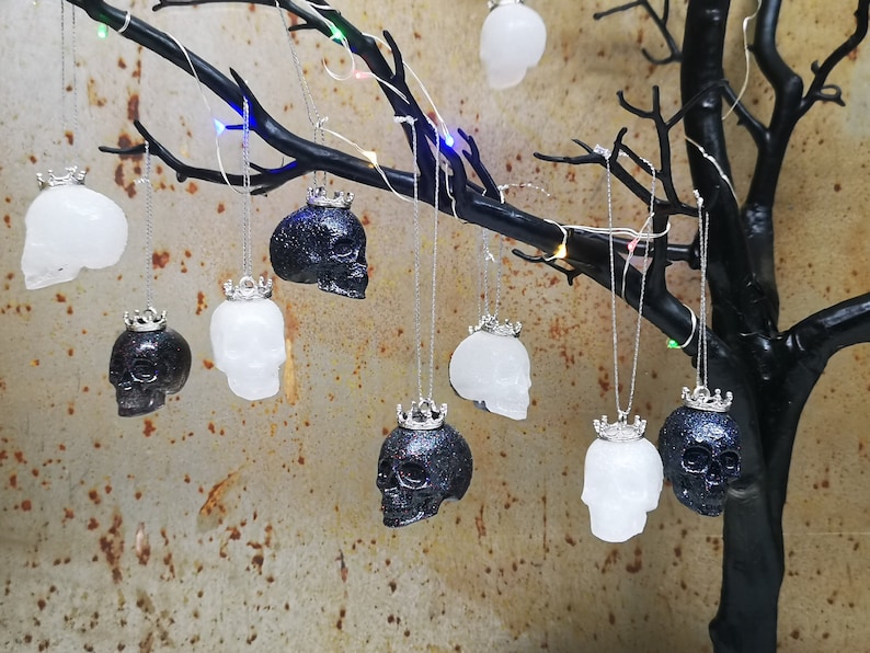 May include: A collection of black and white skull ornaments with silver crowns hanging from a branch. The ornaments are made of resin and have a glittery finish.