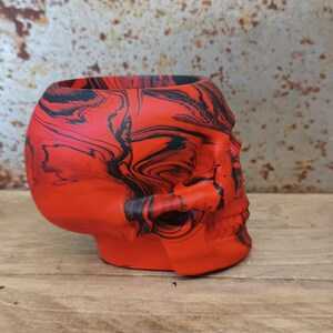 May include: A red and black skull-shaped planter with a marbled design. The planter is made of ceramic and has a smooth, glossy finish.
