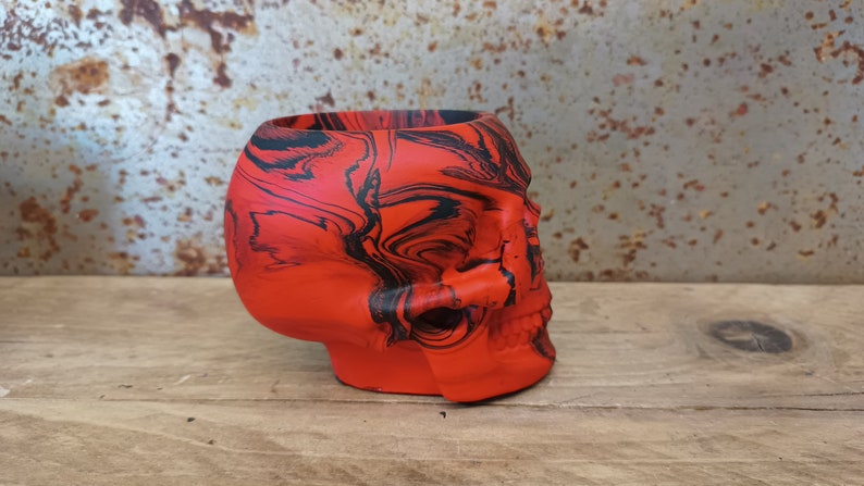 May include: A red and black skull-shaped planter with a marbled design. The planter is made of ceramic and has a smooth, glossy finish.