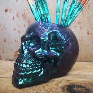 K&ouml;nnte beinhalten: Ein teal- und lilafarbener, schillernder, sch&auml;delf&ouml;rmiger Keramik-Make-up-Pinselhalter mit einem Irokesenschnitt aus teal- und kupferfarbenen Make-up-Pinseln.
