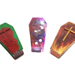 Pu&ograve; includere: Tre scatole in miniatura a forma di bara con una croce dorata in cima. Le scatole sono dipinte in diversi colori: rosso con glitter verde, viola con punti bianchi e marrone con accenti dorati.