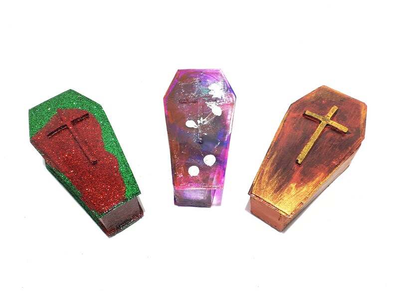 Pu&ograve; includere: Tre scatole in miniatura a forma di bara con una croce dorata in cima. Le scatole sono dipinte in diversi colori: rosso con glitter verde, viola con punti bianchi e marrone con accenti dorati.