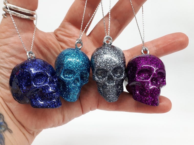 Puede incluir: Cuatro adornos de calavera con purpurina en azul, azul verdoso, plateado y morado. Cada calavera cuelga de una cuerda plateada.